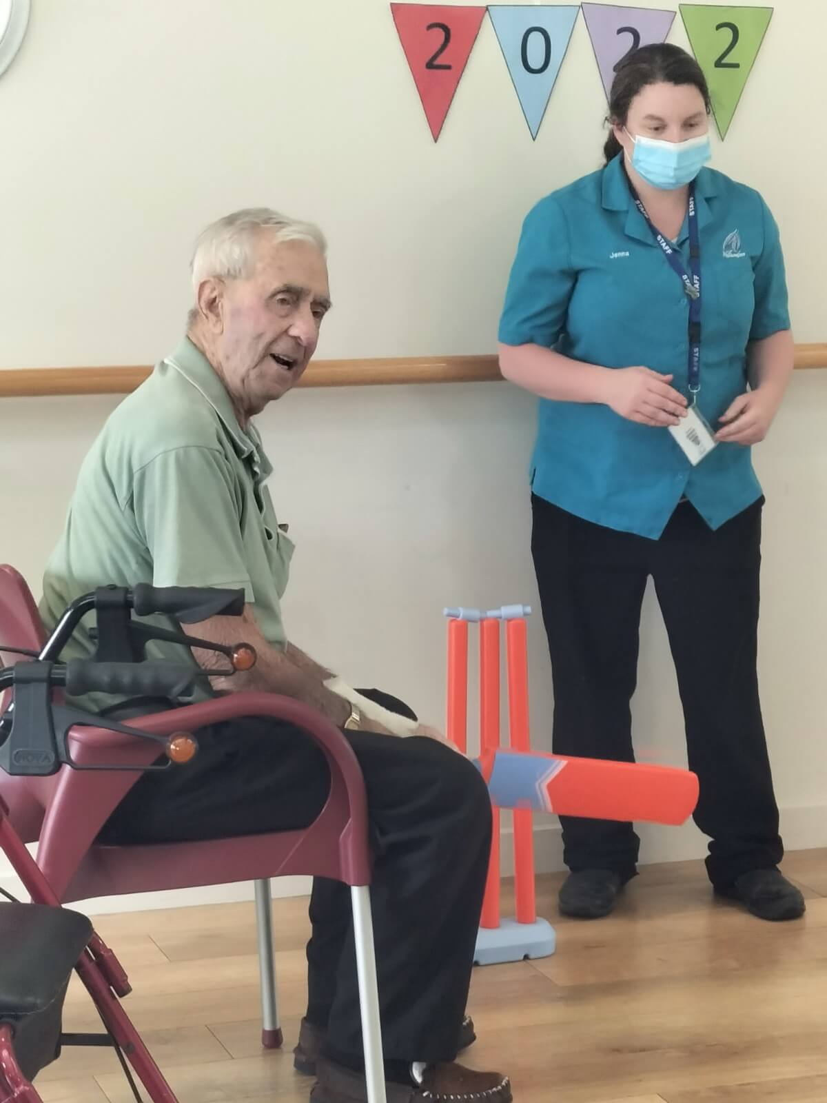 Photo of staff and resident at Yallambee Aged Care as they play indoor cricket. The male residentis holding an orange cricket bat and sitting down while he bats.