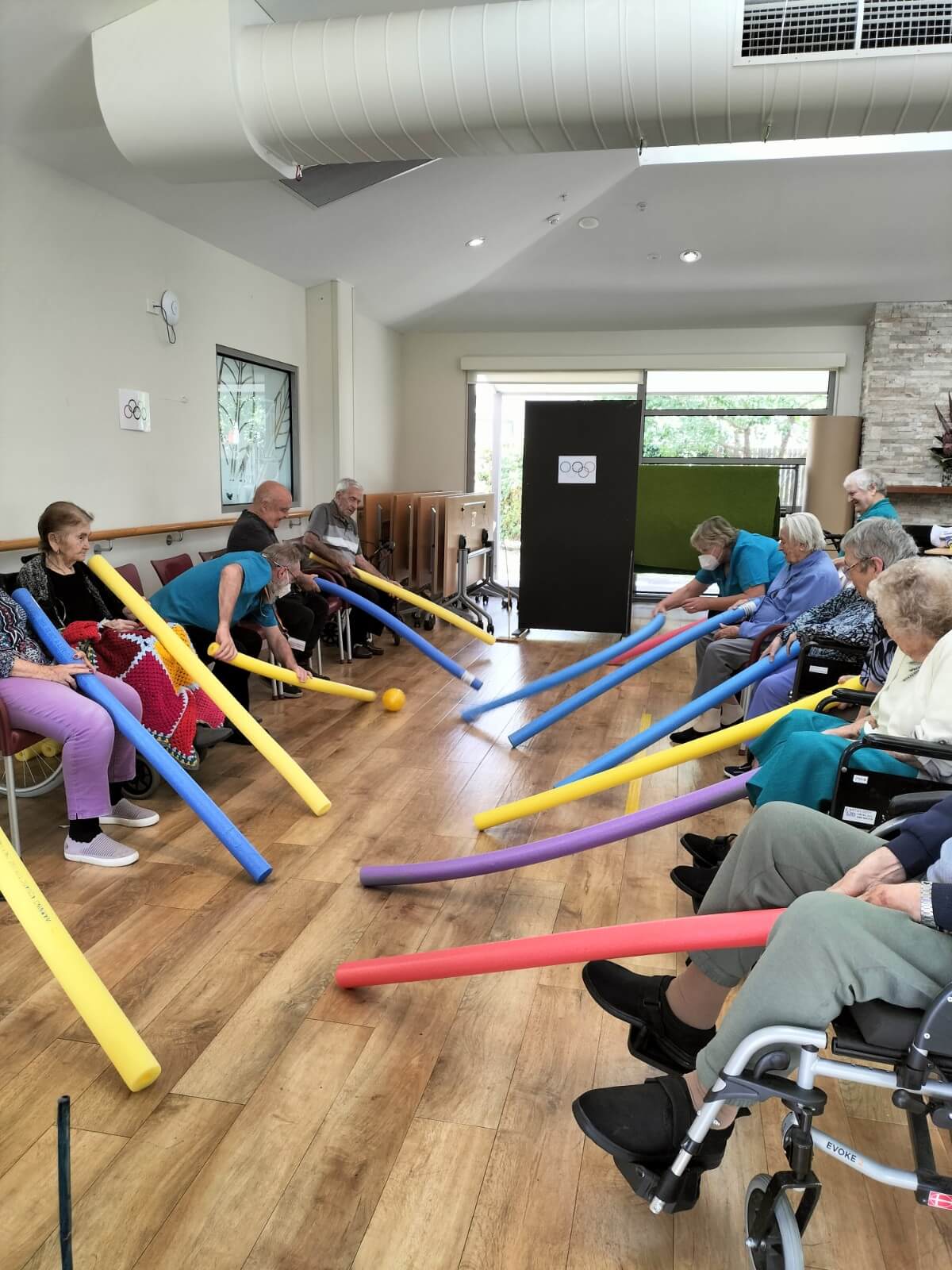 Photo of residents at Yallambee Aged Care sitting down in two rows, playing a game with long foam sticks and trying to push a ball.