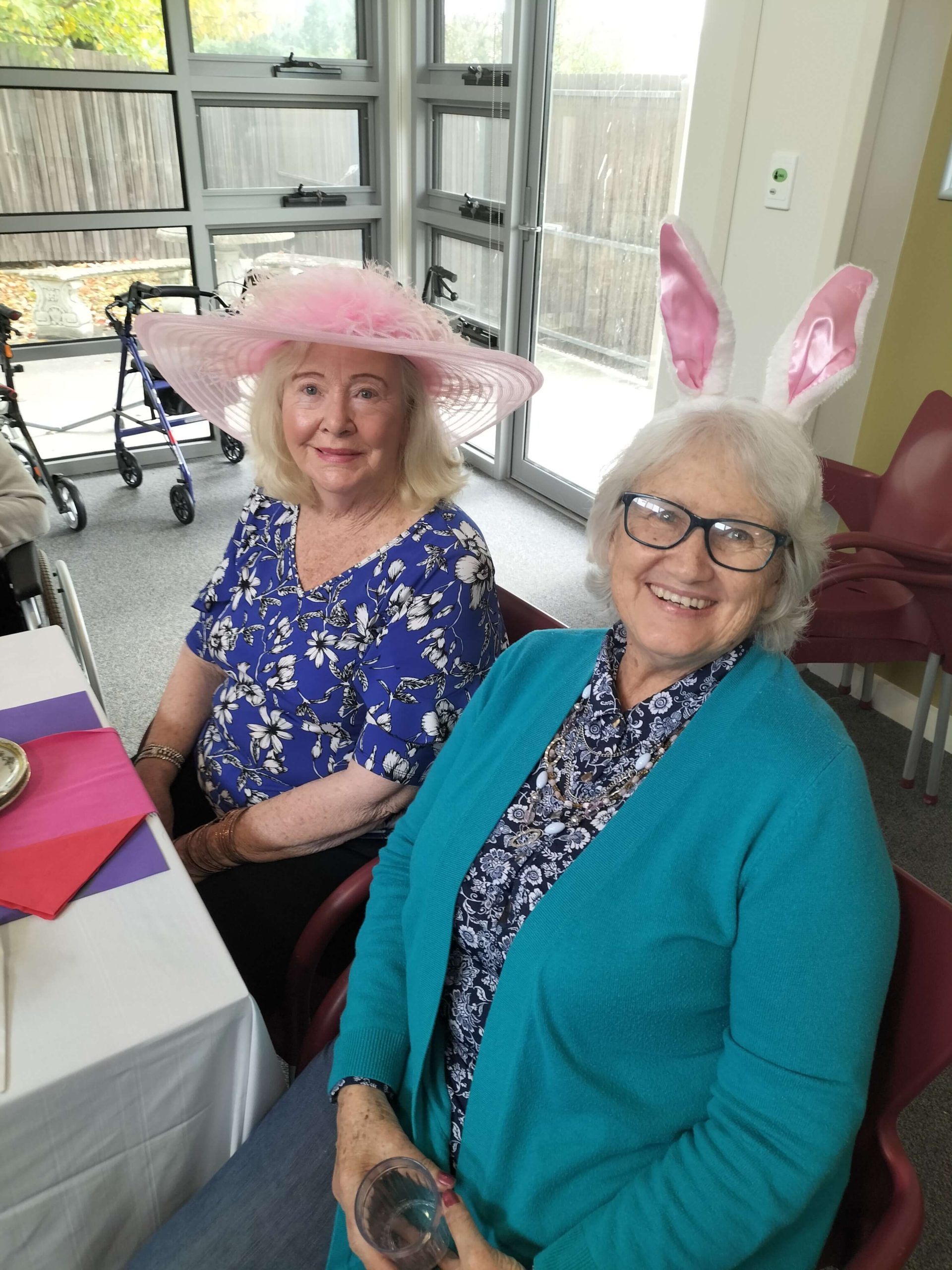 Photo of two elderly ladies sitting down wearing Easter Bonnet hats, one hat is pink with pink feathers on it, the other is pink rabbit ears. Both ladies look like they are having fun.