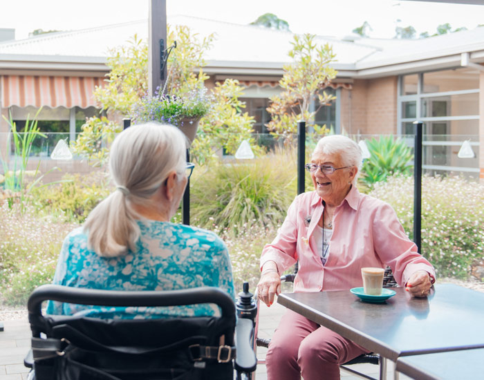 Photo of two elderly ladies sitting in a garden outdoor space, having coffee laughing.