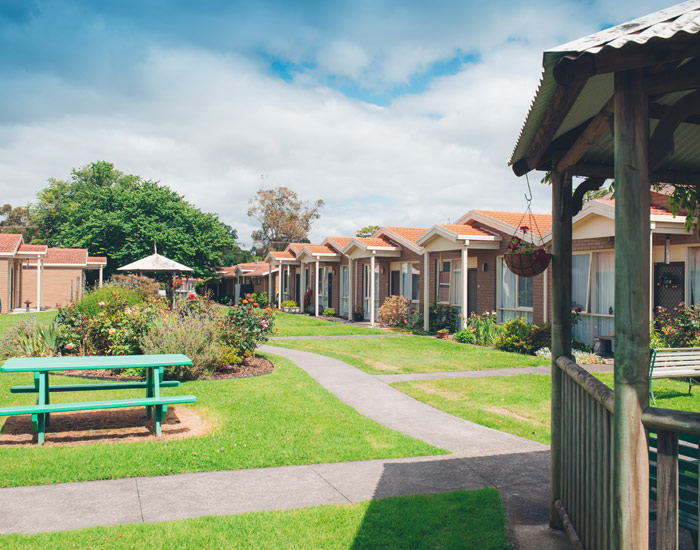 Photo of brown brick units in a row with flower gardens in front of them, some lawn and picnic tables are also infront of the units.
