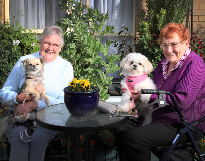 Photo of two elderly ladies and their small dogs as they sit at an outdoor table and chairs in a garden, at Yallambee Aged Care