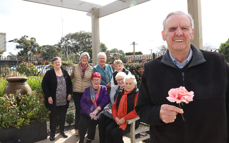 Photo of a group of elderly women sitting and standing in a garden pergola. In the foreground is a man holding a pink rose.