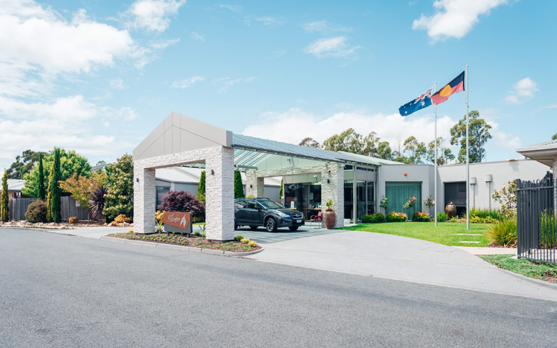 Photo of the driveway entry to Yallambee Aged Care. With a black car parked in white brick entry pavilion. Manicured garden with trees and shrubs, and the Australian and Aboriginal flags flying.