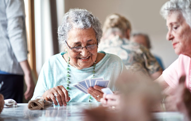 Photo of two elderly women playing cards at a table, with others in the background.