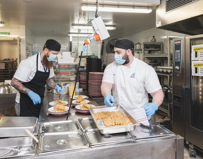 Photo of two chefs working in a commercial kitchen preparing crumbed fish to go into an oven.