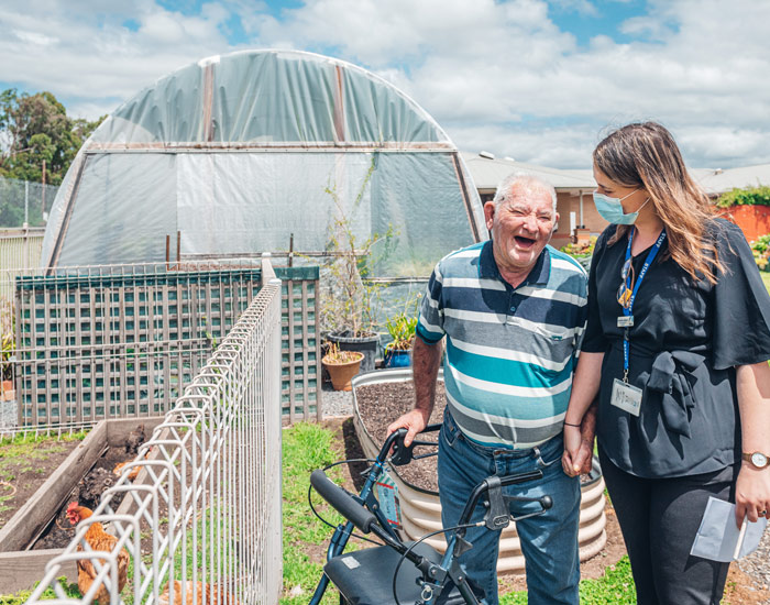 Photo at a nursing home of an elderly man with a walker and a younger women staff member, they are walking through a vegetable garden together and he is laughing.