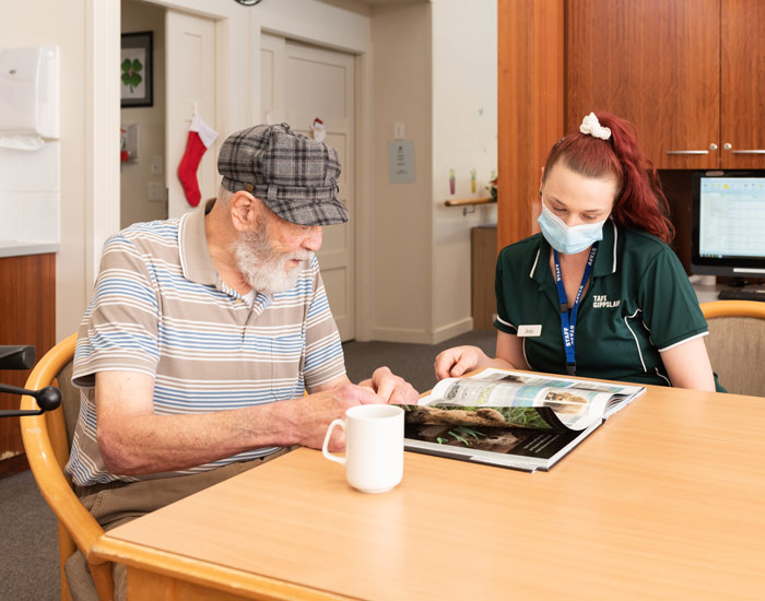 Photo of a nursing home and female staff member is reading a magazine at a table with an elderly man.