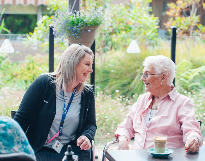 Photo of a young woman sitting down in an outside garden area, in conversation with an older woman. The young woman has blonde hair and wears black, with staff tag on, the older women has grey hair and wears a pink shirt, she has a fresh coffee latte drink.