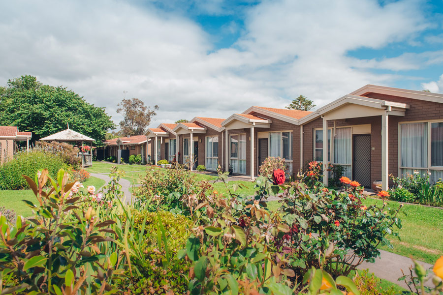 Photo of brown brick units in a row with flower gardens in front of them, some lawn and large trees are also infront of the units.