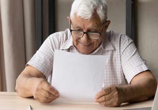 Photo of an elderly man sitting at a table reading a letter, he is smiling and looks happy.